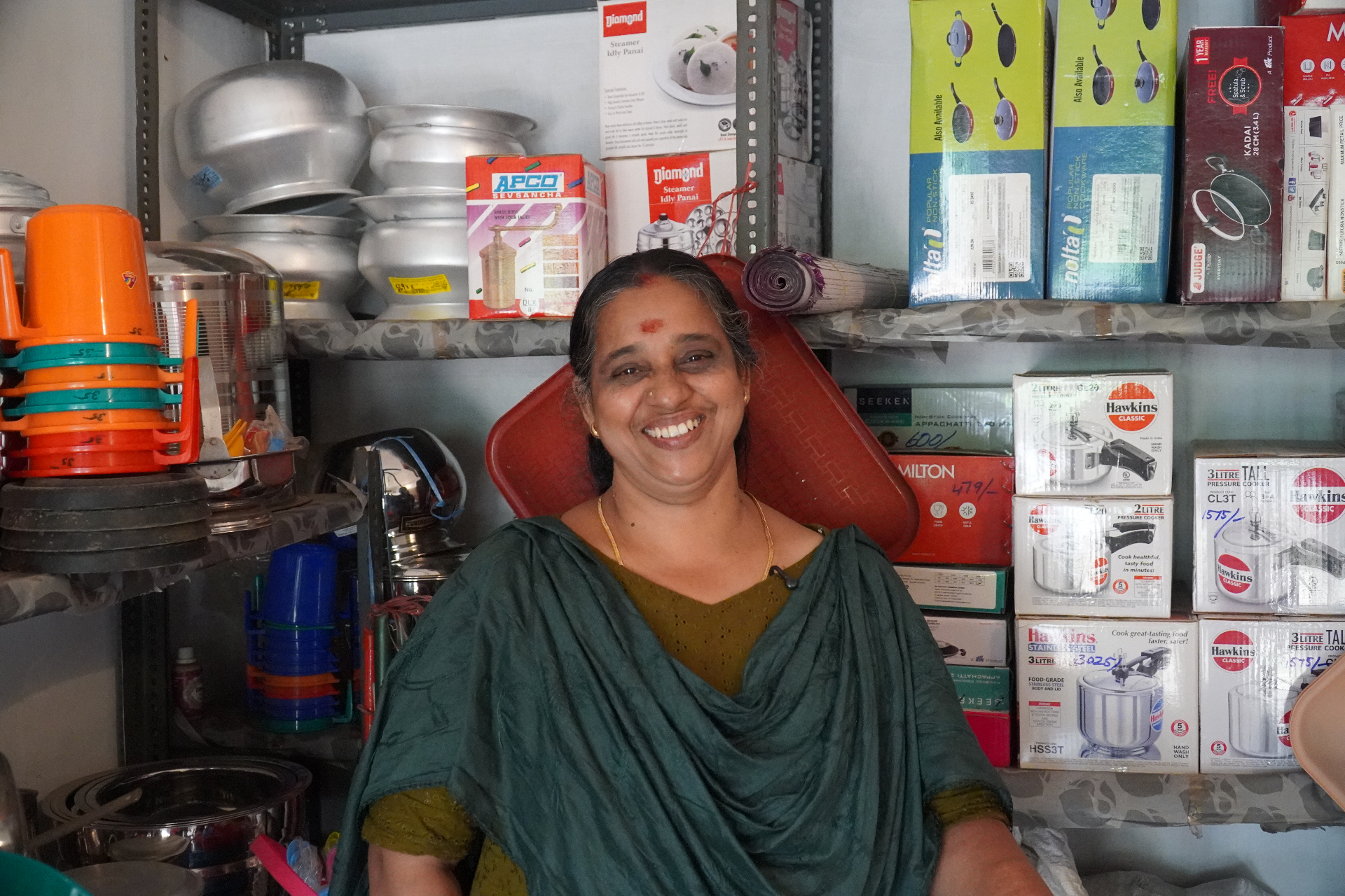 A woman shopkeeper in her Kerala kitchenware store, surrounded by shelves of pressure cookers, idli steamers and stainless steel vessels.