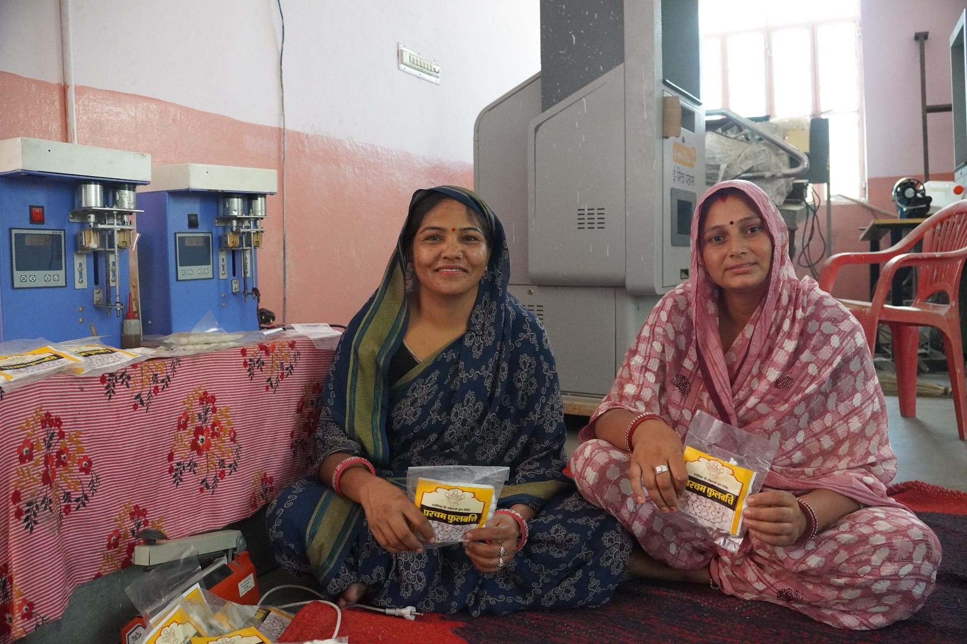 Two women producers at a Rajeevika incense unit in Rajasthan holding packets of Parcham Phulbatti, with pressing machines behind them.