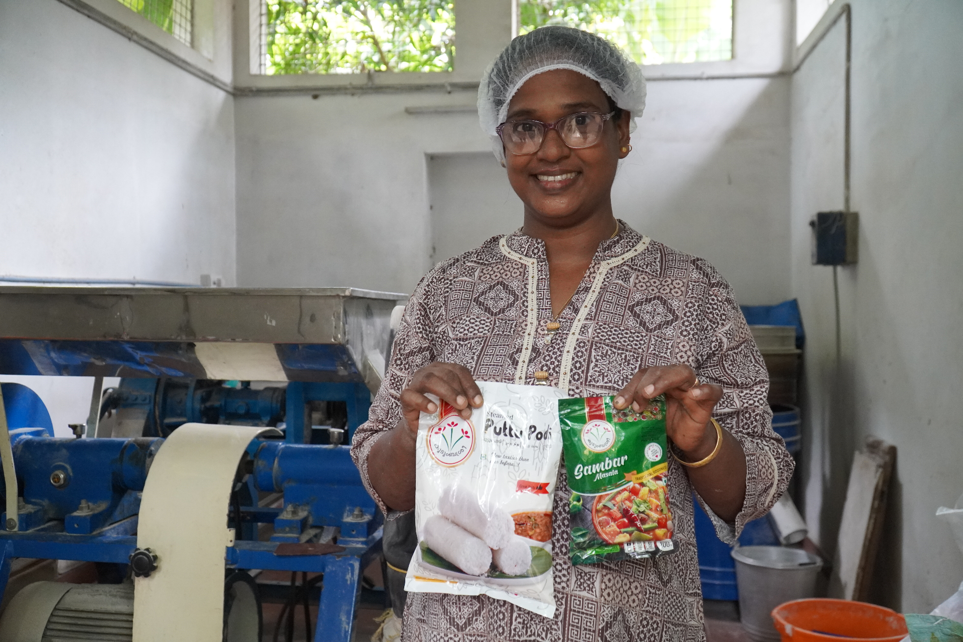 A woman producer at a Kudumbashree food processing unit in Kerala holding packets of puttu podi and sambar masala with a grinder behind her.
